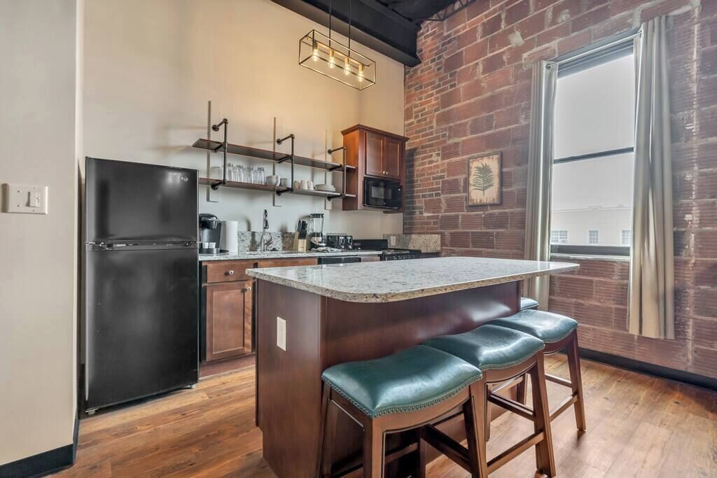 A modern kitchen with a black refrigerator, wooden cabinets, a granite countertop, and a brick wall.