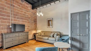 A modern living room with a brick wall, a sofa, a TV on a dresser, and a door.