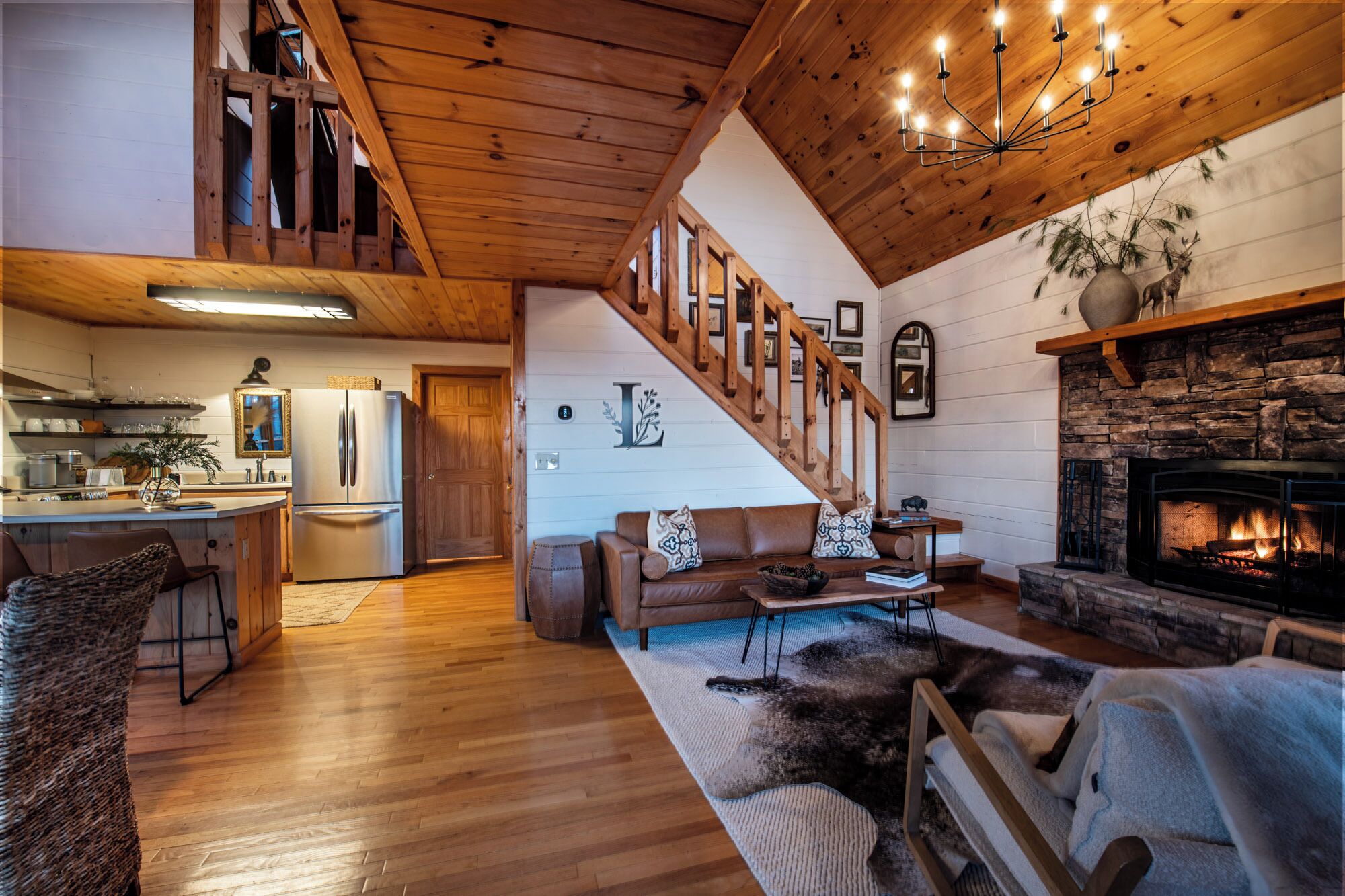A cozy living room with a fireplace, wooden floors, a staircase, and a kitchen area.