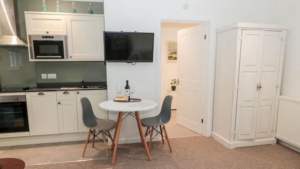 A compact kitchen with white cabinets, a microwave, and a flat-screen TV. There's a small dining table with two chairs and a bottle of wine. A potted plant is visible in the background.