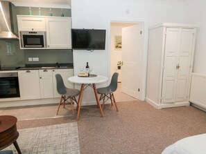 A compact kitchen with white cabinets, a microwave, and a flat-screen TV. There's a small dining table with two chairs and a bottle of wine. A potted plant is visible in the background. - Flodden (Cornhill on Tweed)