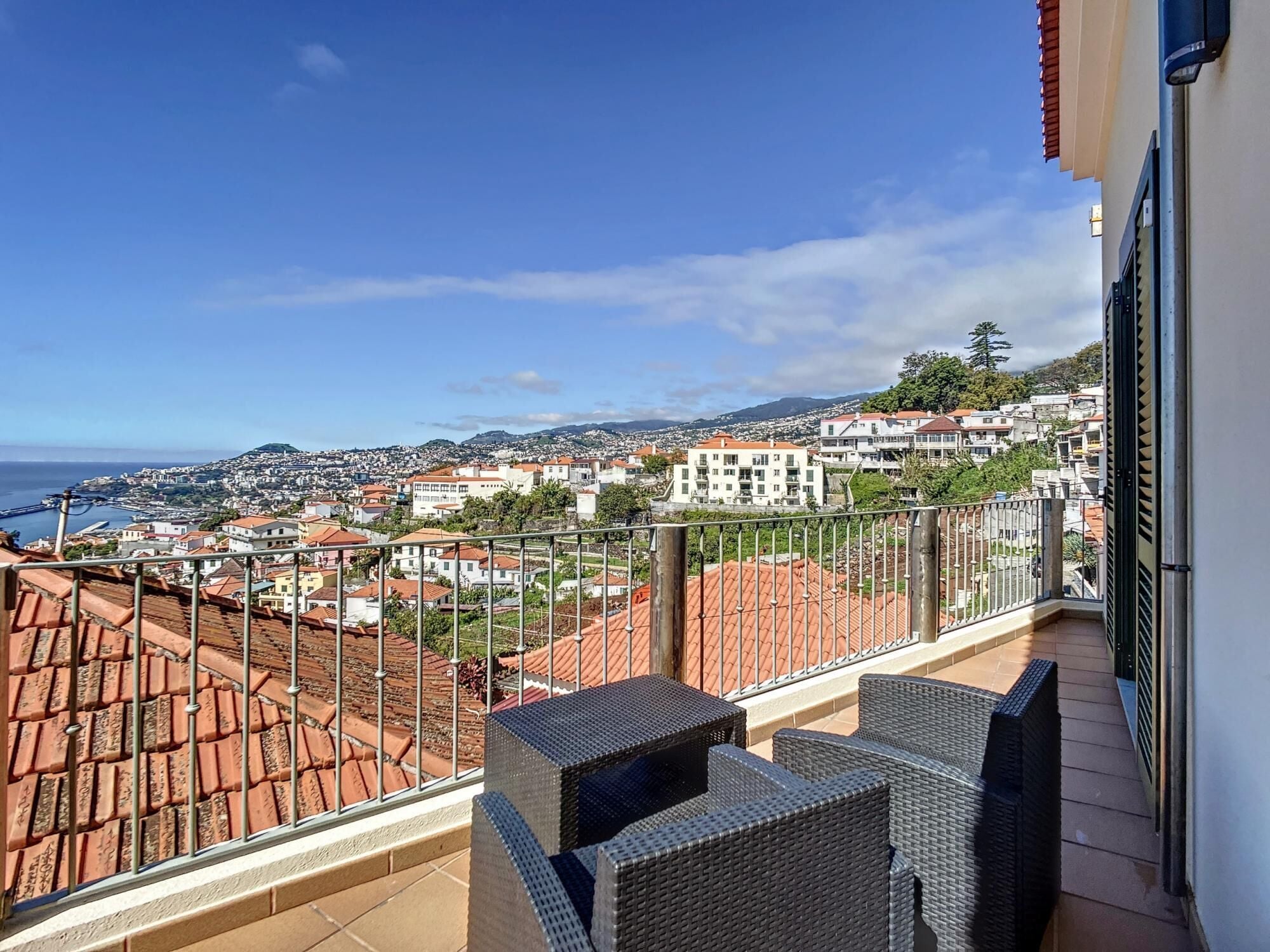 Varanda com móveis de vime com vista para uma paisagem costeira, com telhados de terracota e céu azul claro.