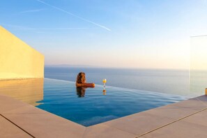 A person relaxing by a clear infinity pool with a glass of drink, overlooking a calm sea. - Syvota 4 Sunset 1B Michaela (Igoumenitsa)