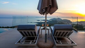 A terrace with two lounge chairs, a table with drinks, and a view of the sea and hills at sunset. - Syvota 08 Sunset 2B Zara (Igoumenitsa)