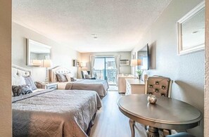 A hotel room with two beds, a round table, a desk, and a balcony with a view.
