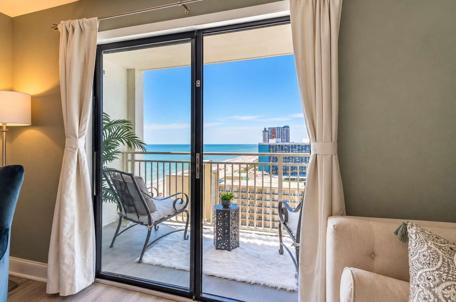 A balcony with a view of the beach and a cityscape, furnished with a chair and a small table.