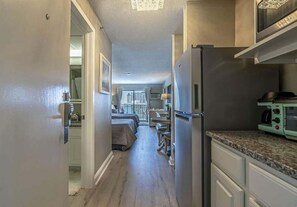 Un pasillo estrecho que conduce a un dormitorio con cama, escritorio y silla. Hay una zona de cocina con refrigerador y microondas.