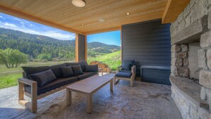 Un espacio de estar al aire libre techado, con chimenea de piedra, muebles de madera y vista a un paisaje boscoso.