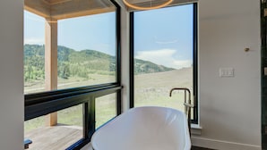 Un baño moderno con una bañera independiente, ventanas amplias que ofrecen vistas a un paisaje boscoso y una terraza de madera en el exterior.
