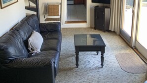 Ein Wohnzimmer mit einem schwarzen Ledersofa, einem Glastisch und einem Deckenventilator aus Holz. Eine Wendeltreppe führt zu einer Küche mit Holzschränken und einer Glastür zum Außenbereich.
