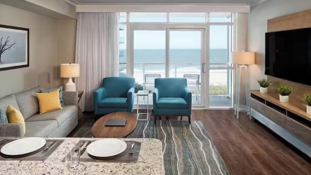 A modern hotel room with a large window offering a view of the sea, a sofa, two armchairs, a coffee table, and a TV on a wooden stand.