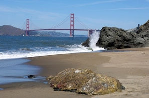 A beach with a large rock in the foreground and a red bridge in the background.
