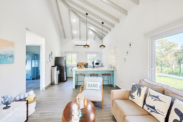 A spacious living room with a high ceiling, a kitchen with a central island, and a large window offering a view of the outdoors.