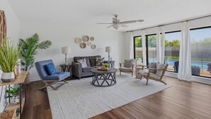 Amplia sala de estar con ventilador de techo, un sofá gris, un sillón azul, una mesa de centro redonda y vista a una piscina a través de grandes ventanales.