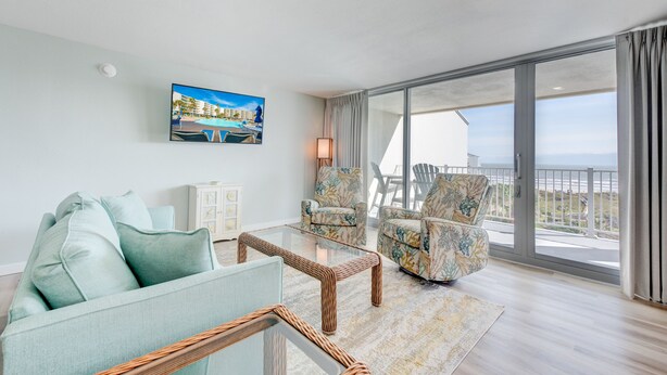 Un salon moderne avec un canapé, des fauteuils et une table basse en verre. Une grande fenêtre offre une vue sur l’océan, et il y a un balcon.