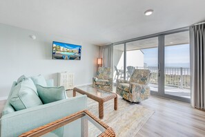 A modern living room with a sofa, armchairs, and a glass coffee table. There is a large window with a view of the ocean and a balcony.