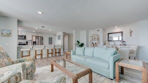 Un salon moderne avec un canapé bleu clair, des tables basses en osier et un coin cuisine à l’arrière-plan.