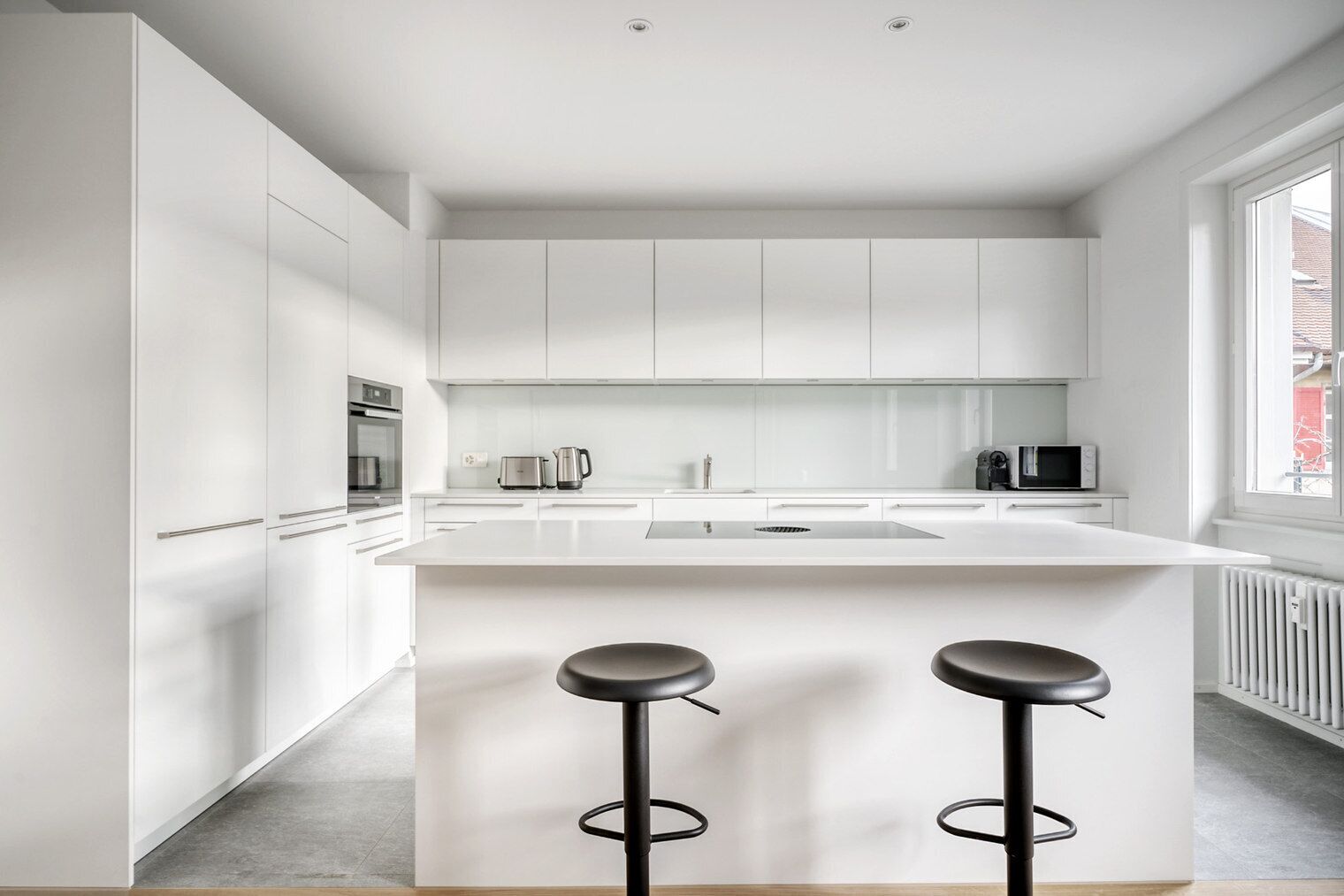 Une cuisine moderne avec des meubles blancs, un îlot central doté de tabourets et une grande fenêtre qui laisse entrer la lumière naturelle.
