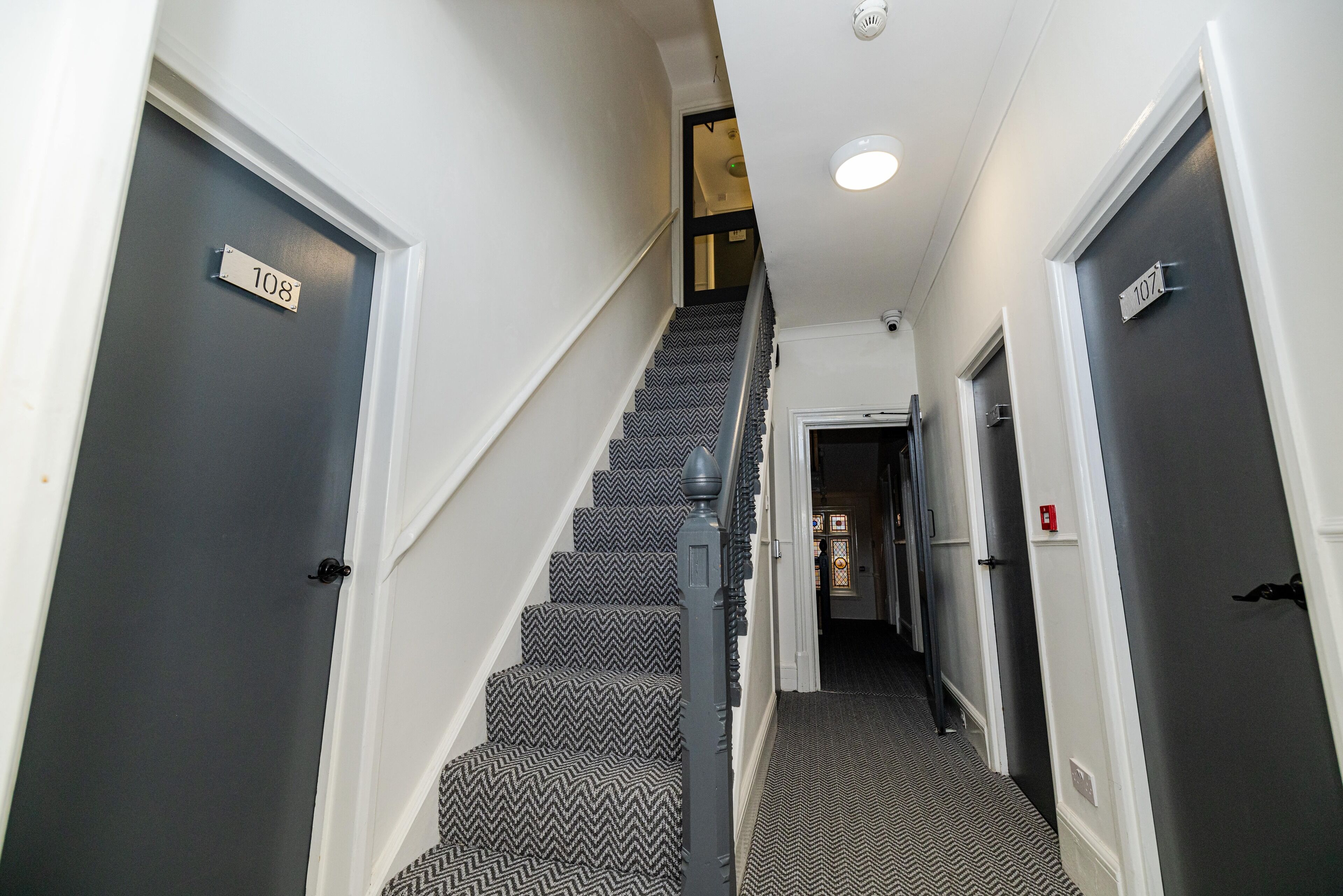 A narrow hallway with a staircase leading to another level. The walls are painted white, and there are door numbers 108 and 107 visible.