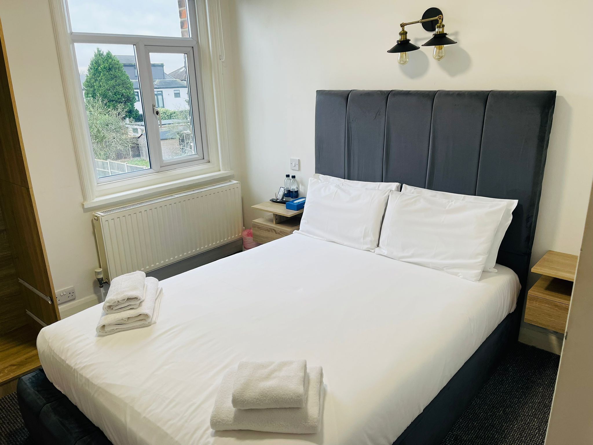 A neatly made bed with white linens and towels, a dark grey headboard, a bedside table with a lamp, and a window with a view of houses and trees.