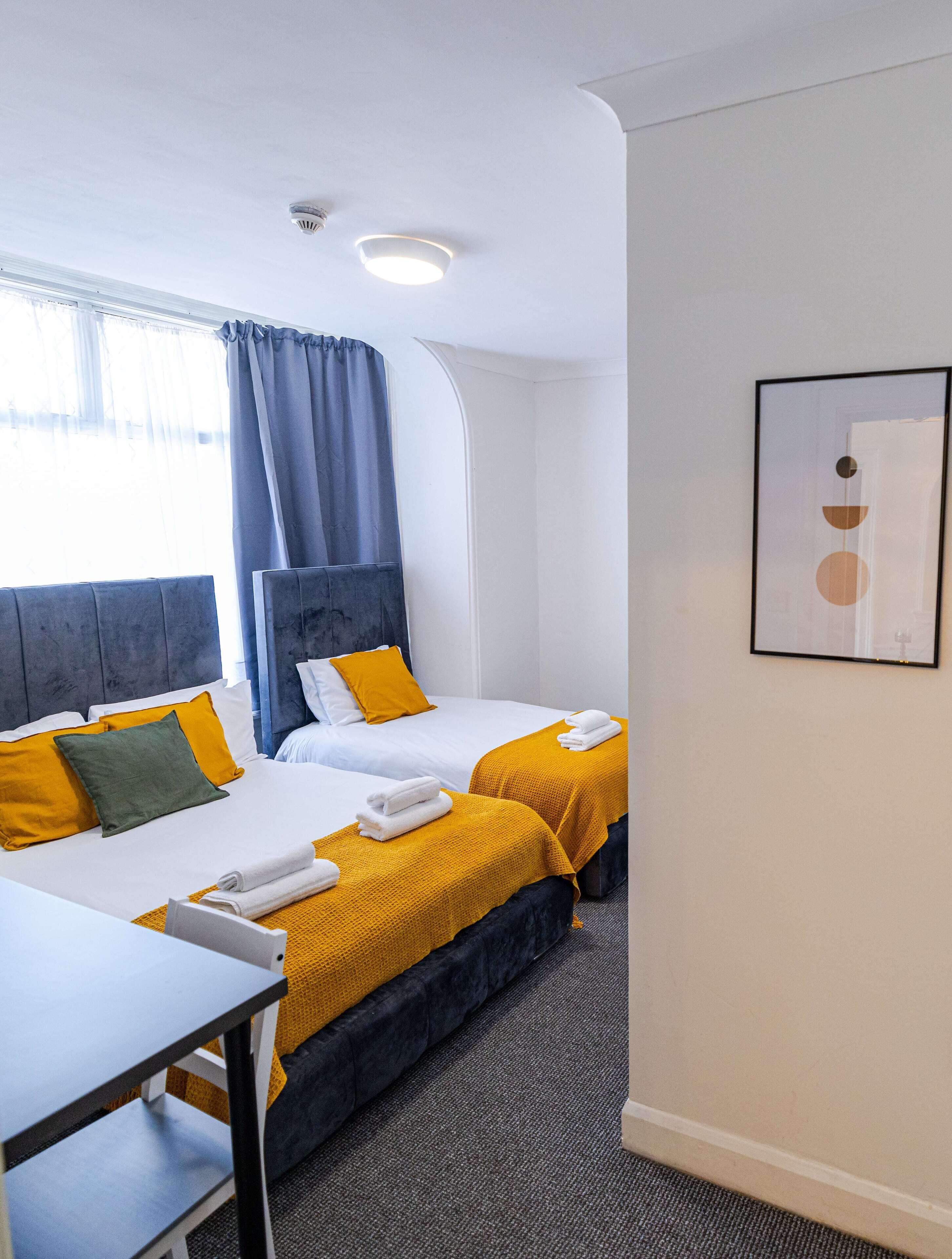 A hotel room with two beds, a desk, a chair, a window with curtains, a ceiling light, and a framed picture on the wall.