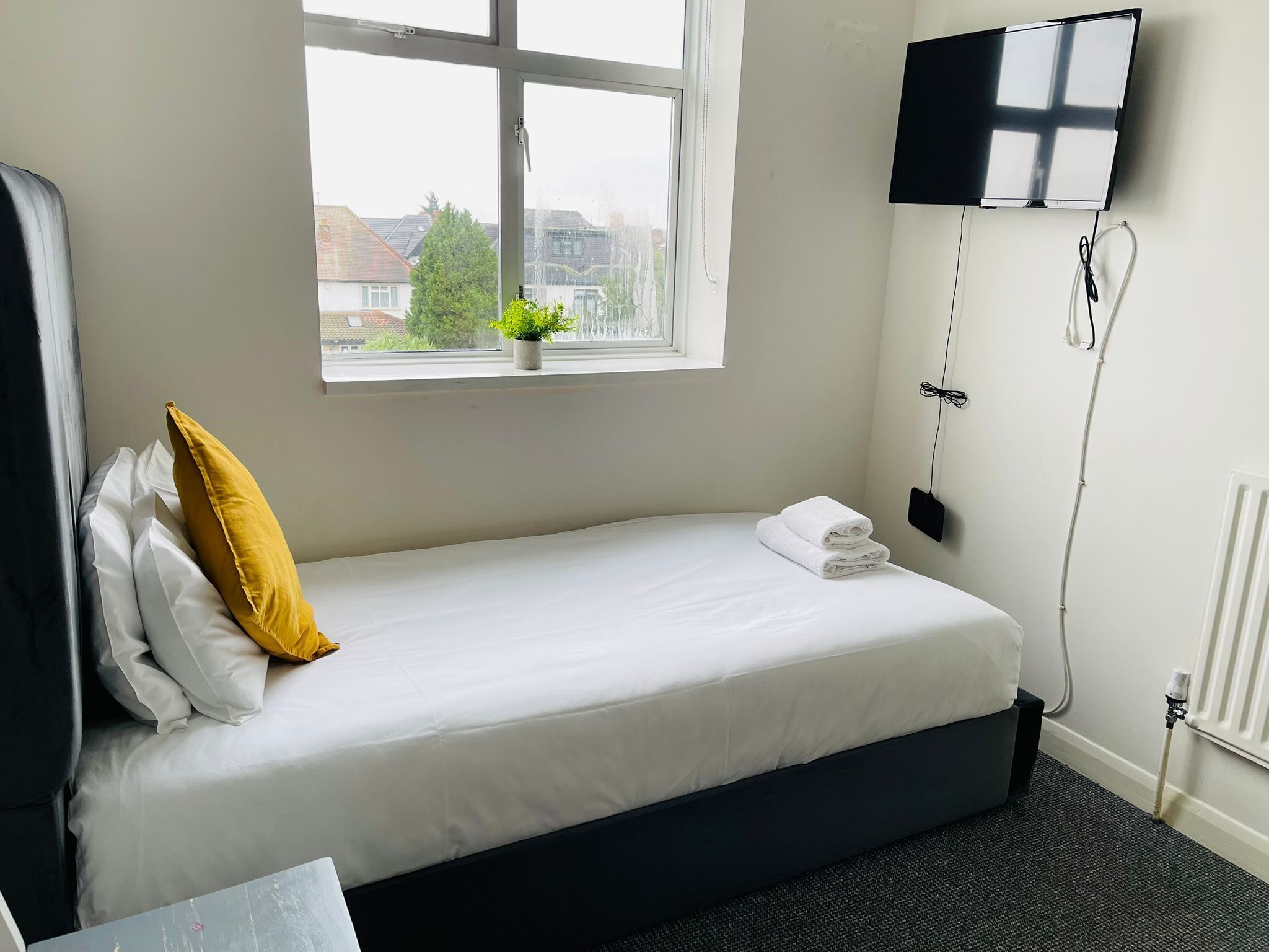 A neatly made bed with white linens and a single yellow pillow, a window with a view of buildings and trees, a flat-screen TV mounted on the wall, and a radiator on the floor.