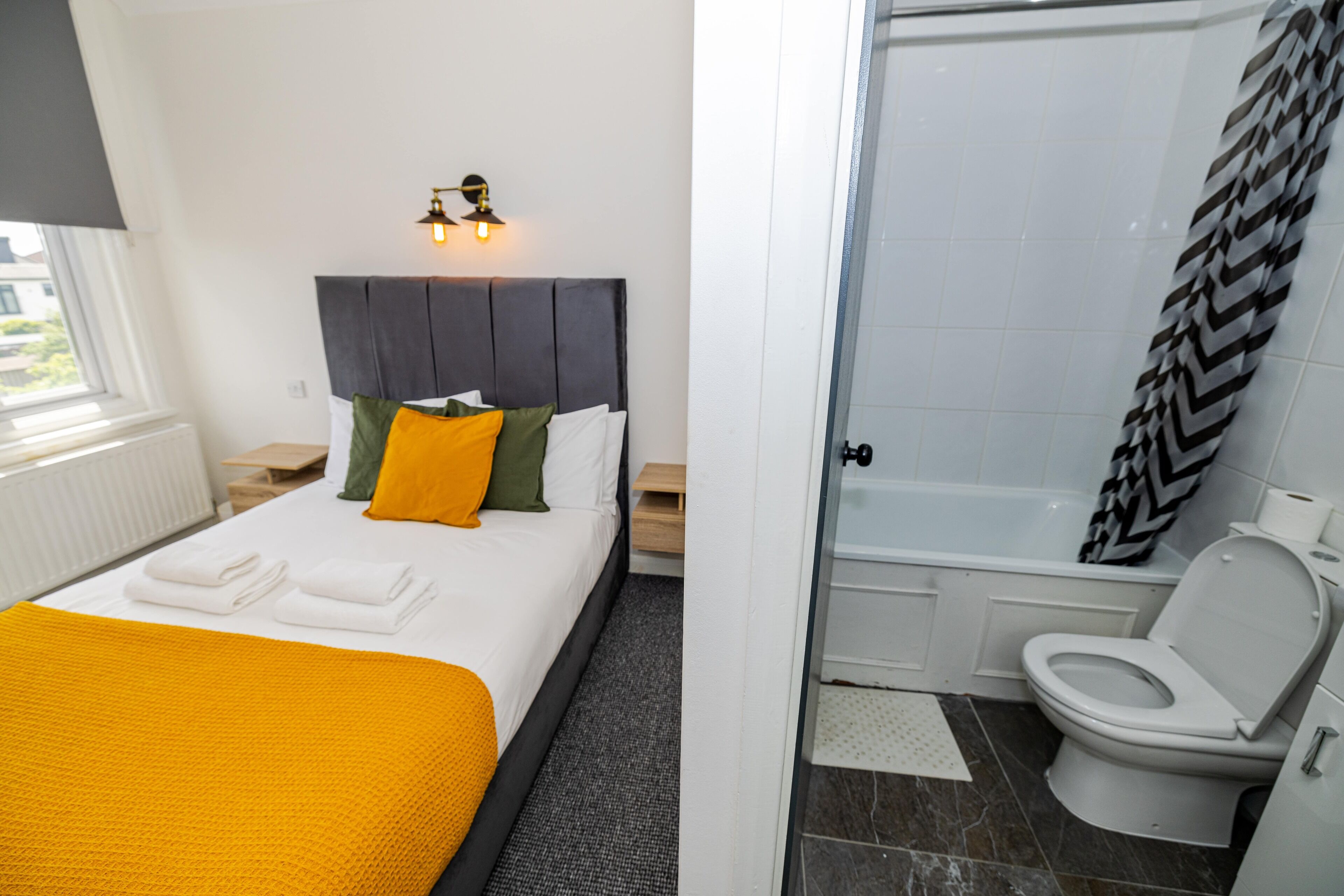 A hotel room with a bed, a nightstand, a lamp, a bathroom with a bathtub, a toilet, and a shower.