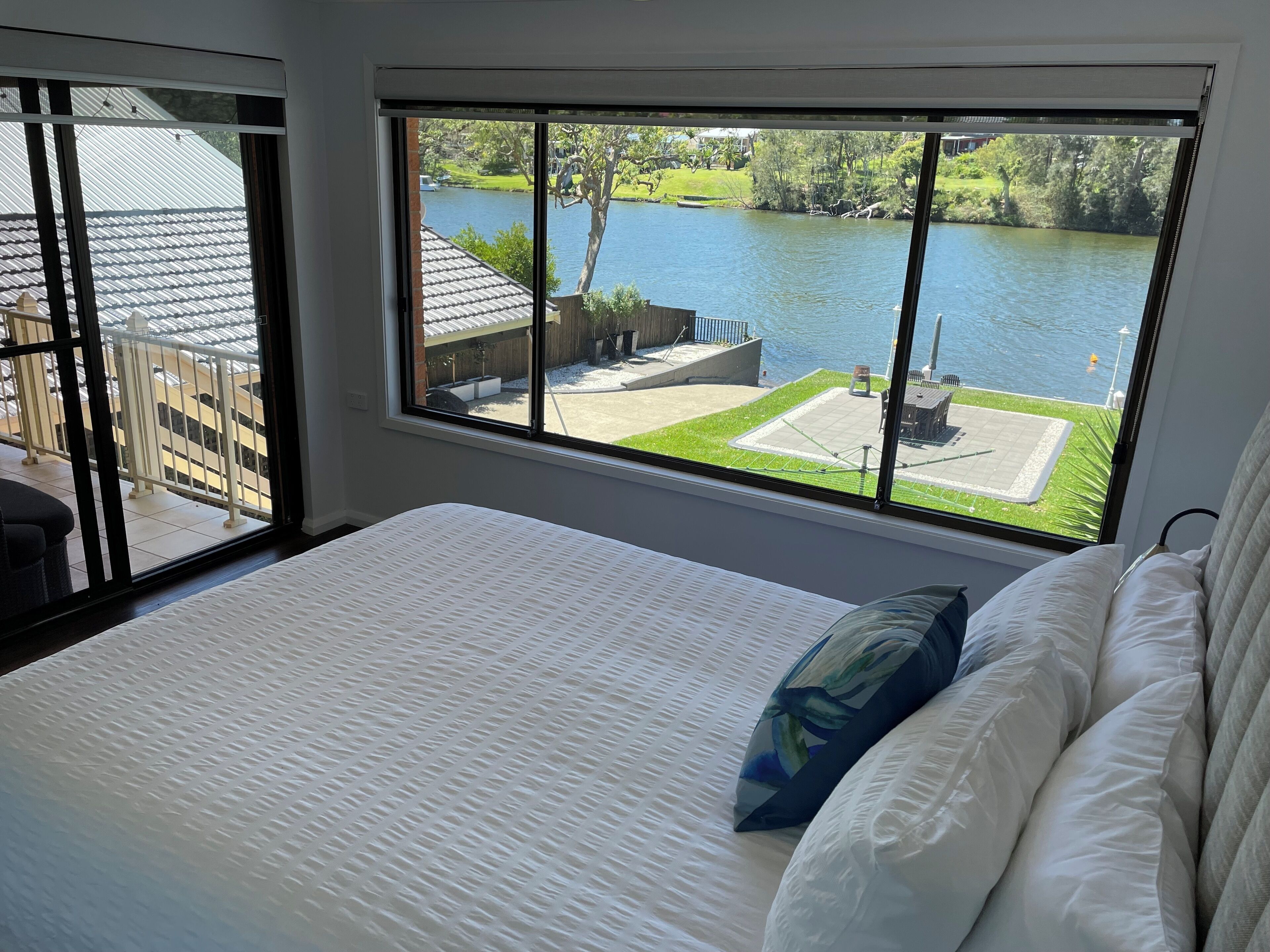 A bedroom with a large window overlooking a lake, a bed with white bedding, and a view of a deck and outdoor furniture.