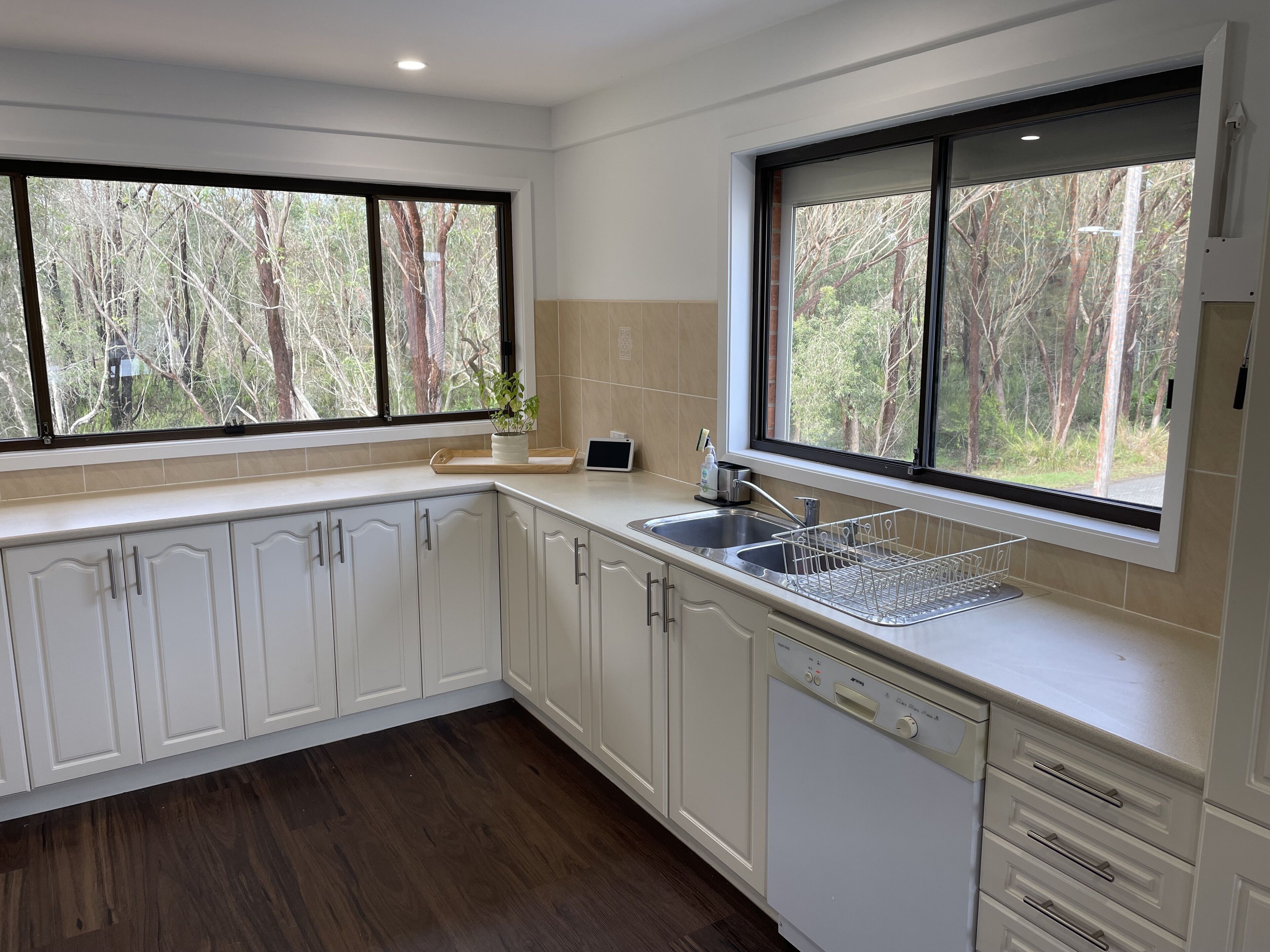 A modern kitchen with white cabinetry, a dishwasher, a sink, and a window overlooking a forest.