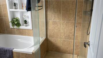A bathroom with a glass-enclosed shower, a white bathtub, and a shelf with plants and toiletries.