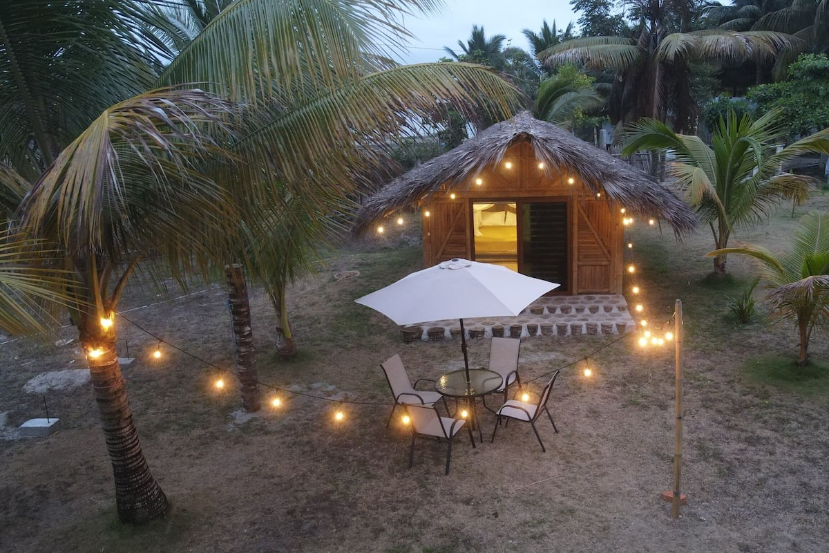 Una cabaña con techo de paja, con una mesa y sillas, rodeada de palmeras y luces colgantes.
