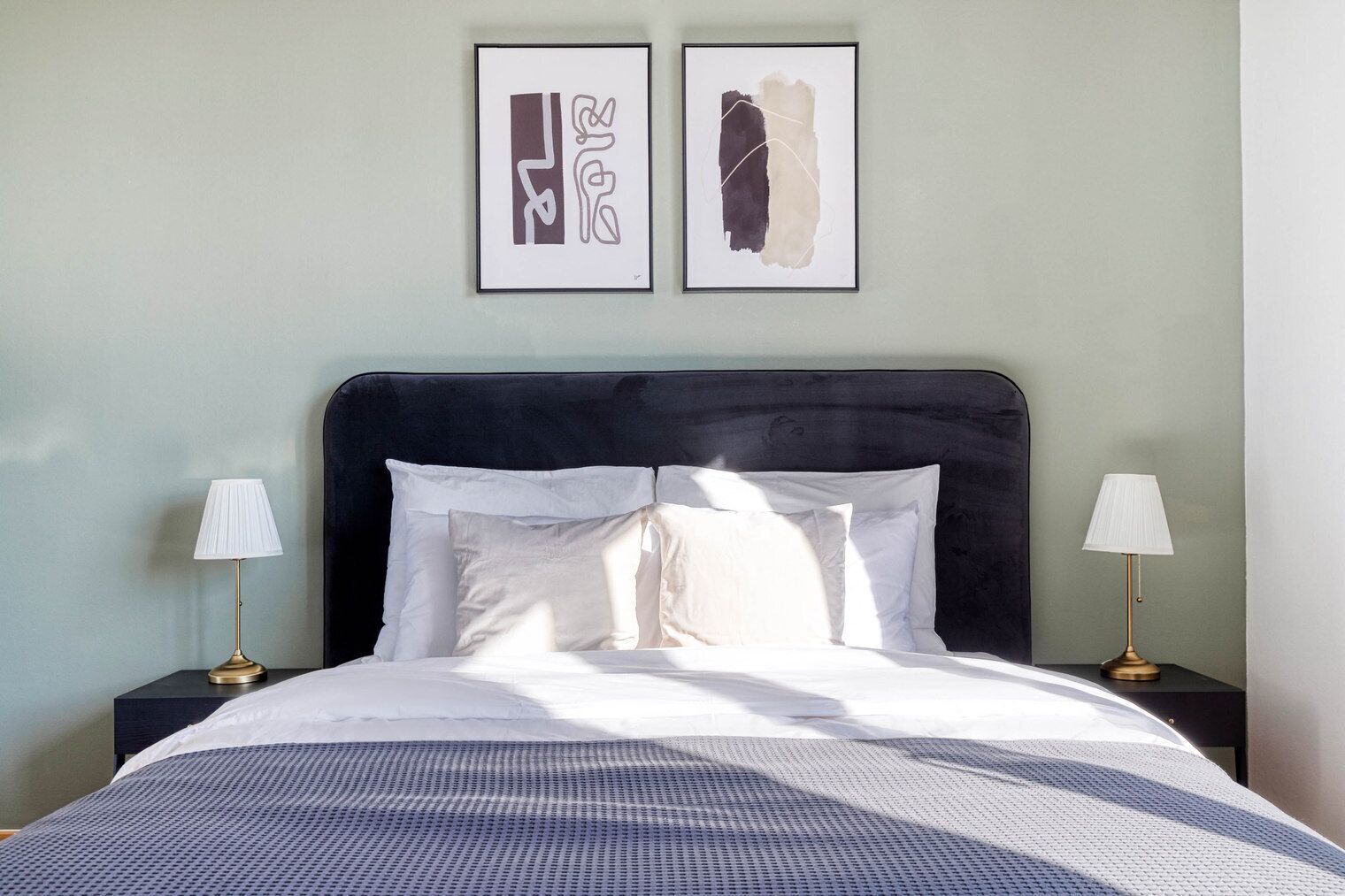 A neatly made bed with white linens and a blue patterned bedspread, flanked by two bedside tables with lamps, against a light green wall.