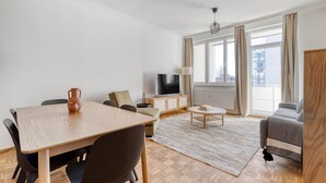 A modern living room with a wooden dining table, a sofa, a TV, and a large window with curtains. - Refurbished Penzing 3BR w/ Elevator & Balcony, nr metro U3, by Blueground (Vienna)
