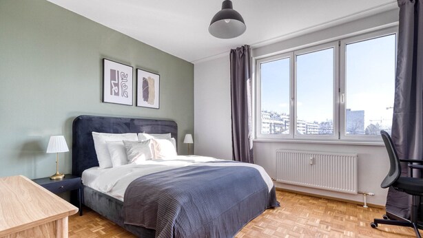 A modern hotel room with a large bed, a desk, a chair, and a view of the city through the window. - Refurbished Penzing 3BR w/ Elevator & Balcony, nr metro U3, by Blueground (Vienna)