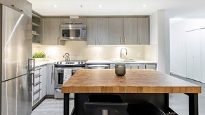 A modern kitchen with stainless steel appliances, grey cabinets, and a wooden dining table. - New Modern, Large 1 Bedroom, 1 Bathroom + Work Space (Vancouver)