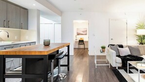 A modern kitchen with a wooden table and bar stools, a white sofa, and a small side table with a lamp. - New Modern, Large 1 Bedroom, 1 Bathroom + Work Space (Vancouver)