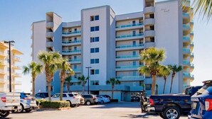 Un hôtel à plusieurs étages au design moderne, doté de balcons à chaque niveau, d’un parking où sont stationnées plusieurs voitures et de palmiers à l’avant.