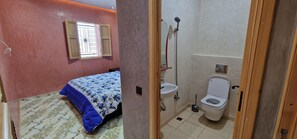 A bedroom with a bed, a window with wooden shutters, and a bathroom with a toilet and sink.