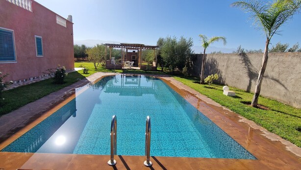 Una piscina con pérgola y zona de estar exterior, rodeada de un césped bien cuidado y un edificio con amplios ventanales.