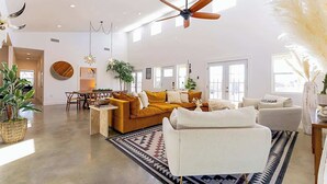 A spacious living room with a large sofa, a coffee table, and a dining area with chairs. There is a ceiling fan, large windows, and a variety of plants. - Sleeps 18, Ranch - Pool, Hot Tub, 2 Fire Pits (Dripping Springs)