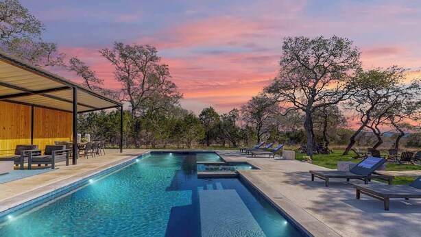 A swimming pool with lounge chairs and a covered seating area, surrounded by trees and a sunset sky. - Sleeps 18, Ranch - Pool, Hot Tub, 2 Fire Pits (Dripping Springs)