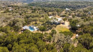 An aerial view of a property with a swimming pool, surrounded by dense vegetation and a few buildings. - Sleeps 18, Ranch - Pool, Hot Tub, 2 Fire Pits (Dripping Springs)