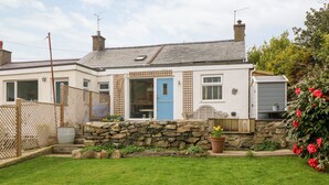 Une maison à deux étages avec un mur en pierre, une porte bleue et un jardin comprenant une pelouse et des plantes en pot.