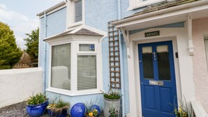 Une maison bleue avec une porte bleue, une fenêtre avec des stores et plusieurs plantes en pot devant.