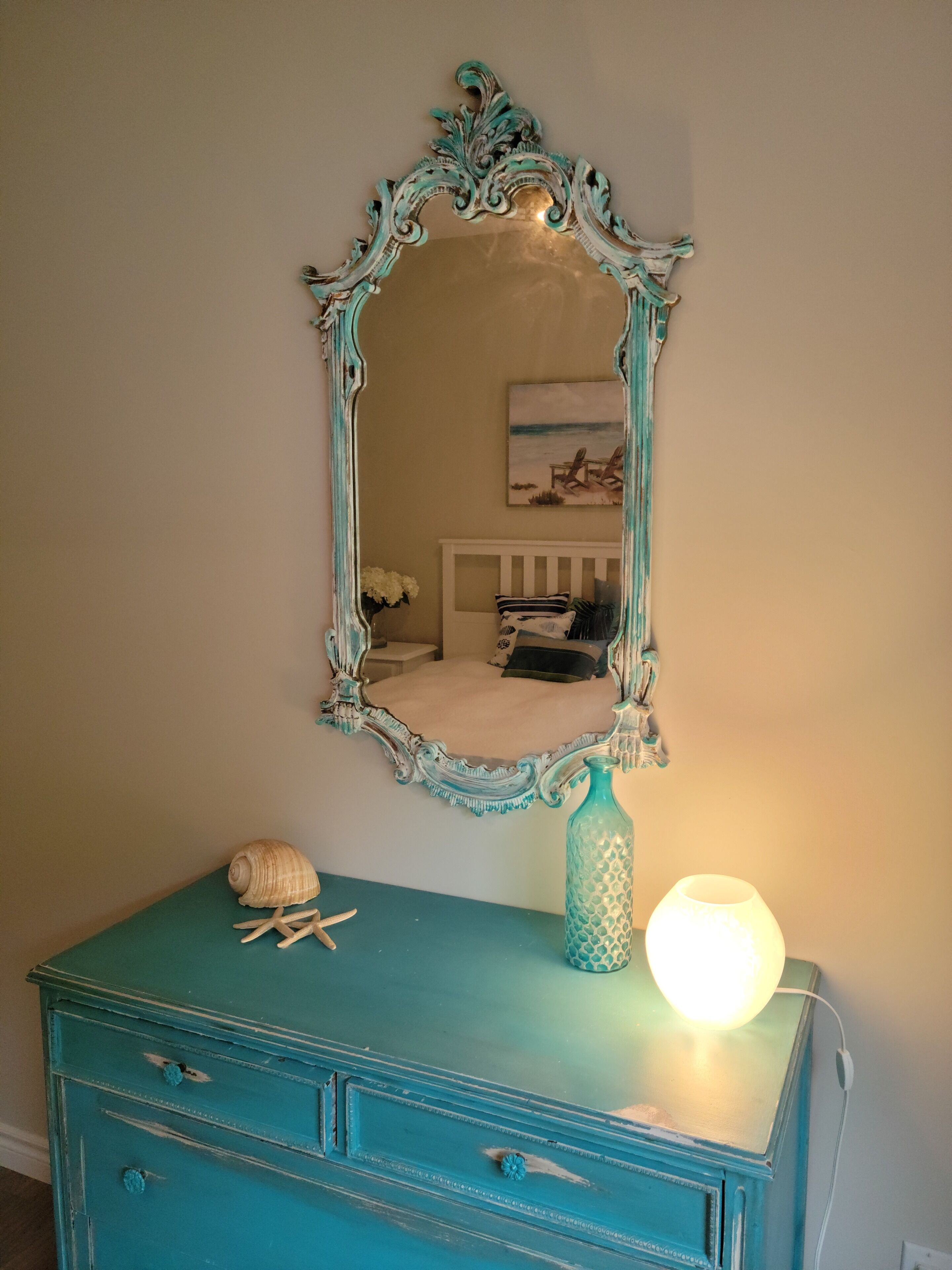 A teal dresser with a decorative mirror above it, a vase, and a lamp on the dresser.