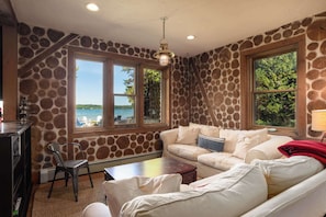 Una habitación con pared de piedra, un sofá blanco, una silla, una mesa y ventanas amplias con vistas al exterior.