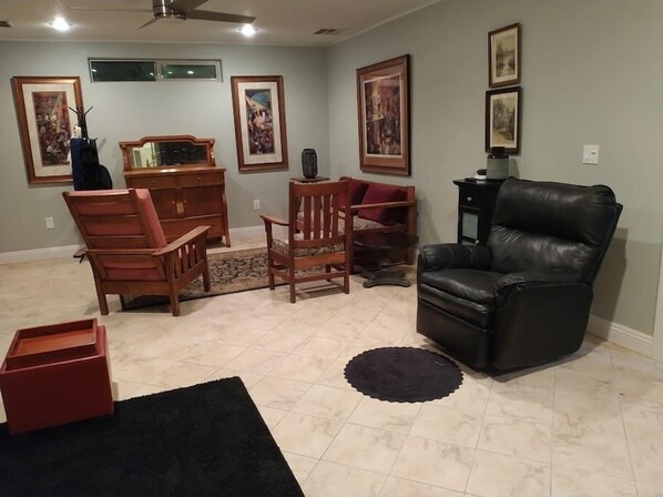 A living room with a black leather chair, wooden chairs, a small table, and a cabinet. There are framed pictures on the wall and a ceiling fan. - Studio in Pahrump | Death Valley NP (Pahrump)