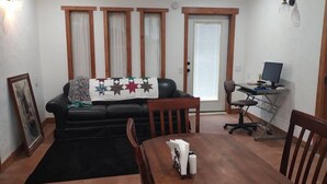 A living room with a black leather sofa, a wooden dining table with chairs, a computer desk, and a large window with curtains. - Studio in Pahrump | Death Valley NP (Pahrump)