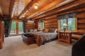 A log cabin bedroom with a bed, bedside tables, a dresser, and a view of the forest through large windows.