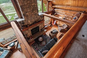 A log cabin interior with a stone fireplace, wooden furniture, and a view of the outdoors.